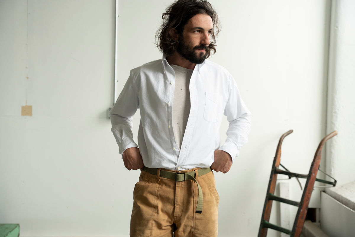 A man with a beard and long dark hair stands indoors wearing a Made in USA white button-down shirt over a light undershirt, tan pants, and a green belt. Ralph tucked styling.  The background is a plain white wall with a wooden ladder visible on the right.