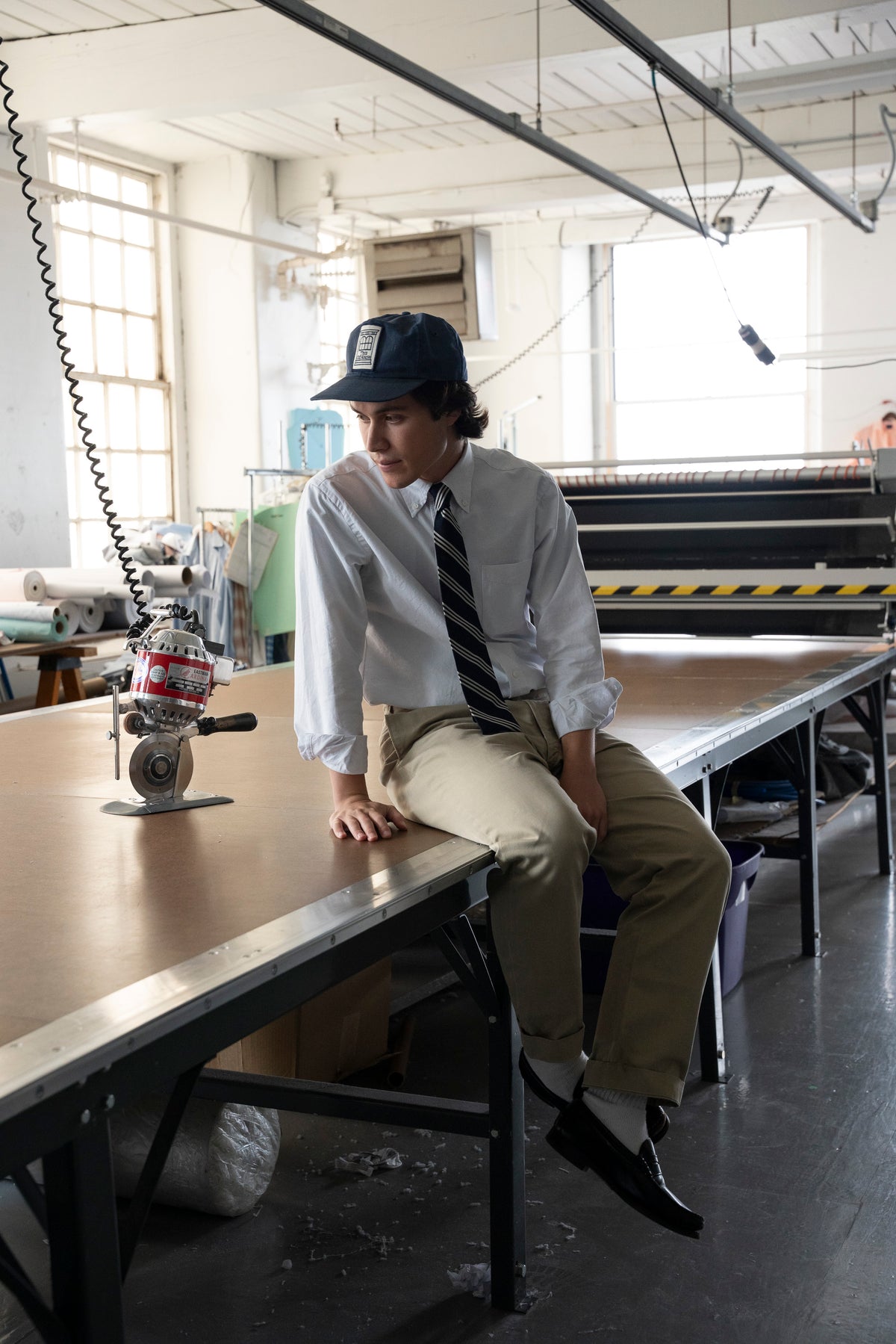 A man wearing a white long-sleeve shirt, striped tie, khaki pants, and a dark cap sits on the edge of a large worktable inside a bright, industrial workshop.  Made in USA Oxford Cloth Button Down factory.