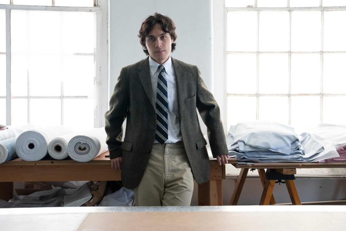 A man in a brown tweed blazer, white Oxford Cloth Button Down shirt, made in USA and sewn in Fall River, MA.  Shot in working factory setting with windows, tables, and fabric rolls in the background.
