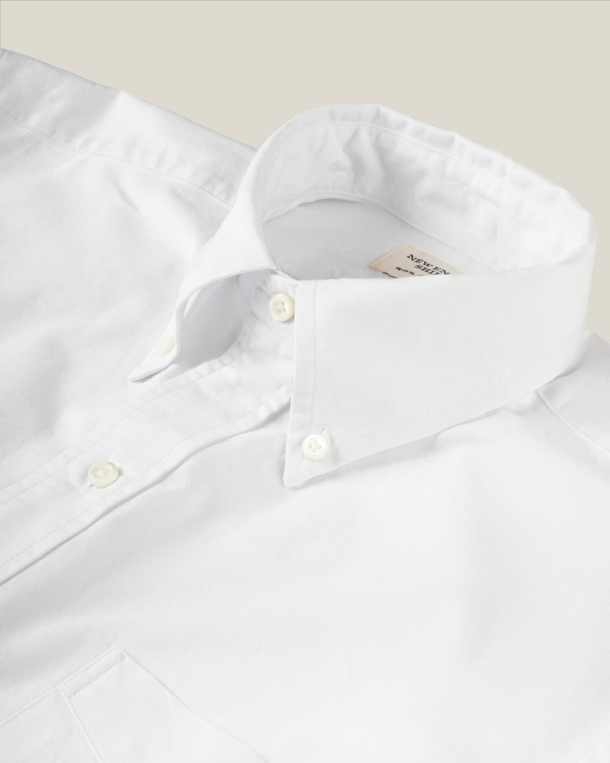 Close-up of a white button-down shirt collar with white buttons, showing part of the inner neck label, set against a light beige background.  Made in USA shirts.