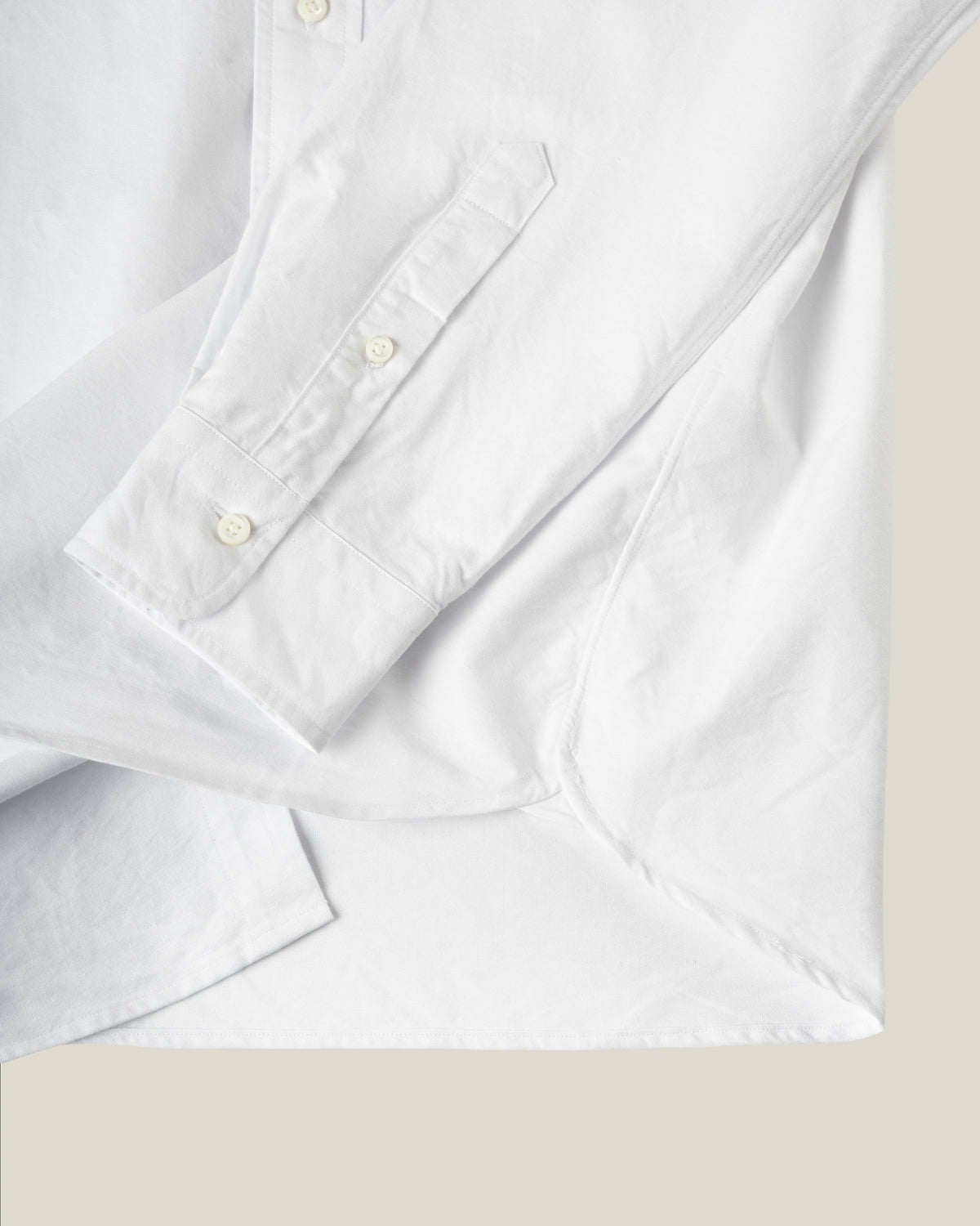 Close-up of a white long-sleeve button-up shirt showing the cuff, buttons, and fabric texture against a light beige background.  Made in USA, sewn in Fall River.