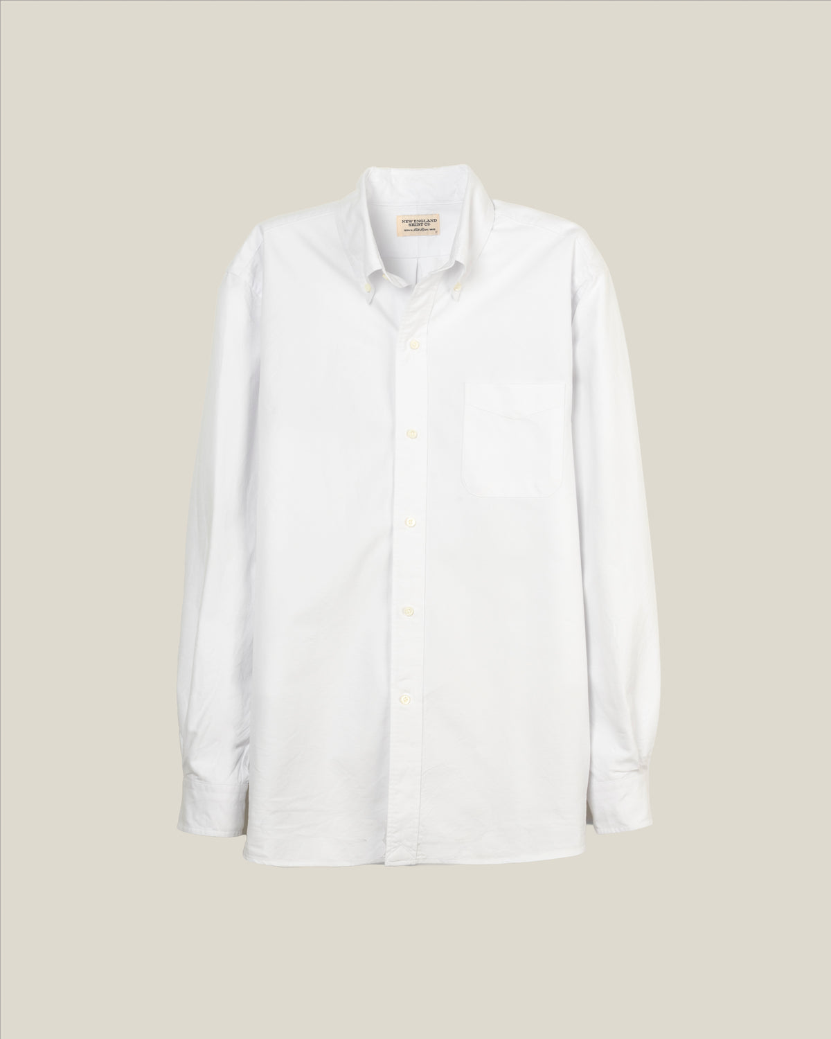 A white long-sleeve button-down shirt with a single chest pocket, displayed flat against a solid light beige background.  Made in USA White OCBD